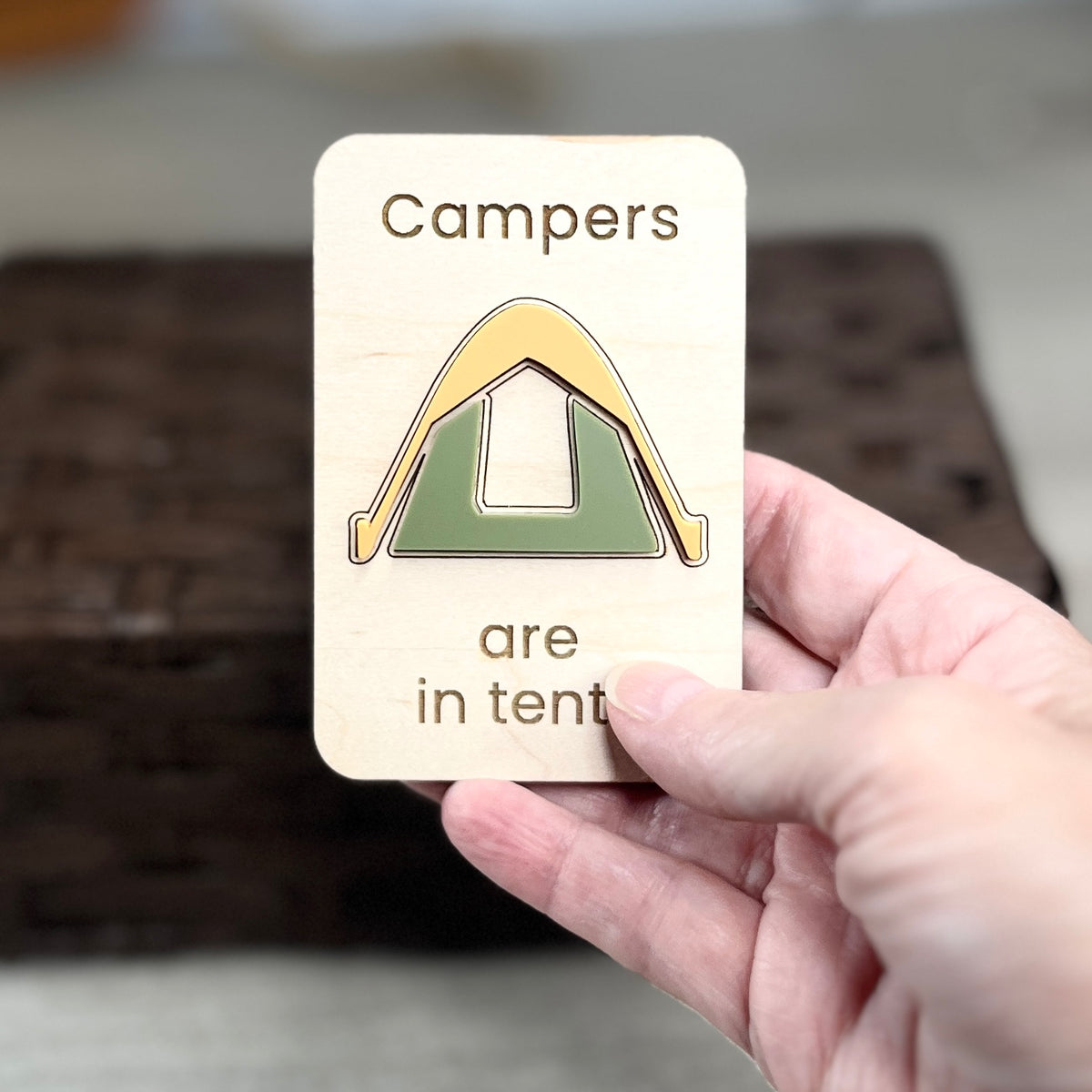 A hand holds a small wooden magnet with the laser-engraved message 'Campers are in tents' and a simple acrylic tent design. The background is softly blurred, showing a dark woven basket and light floor.
