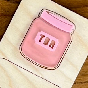 Close up of a bookmark showing a pink acrylic jar shaped like a mason jar with the letters 'TBR' cut out on the front, displayed over a wooden surface.