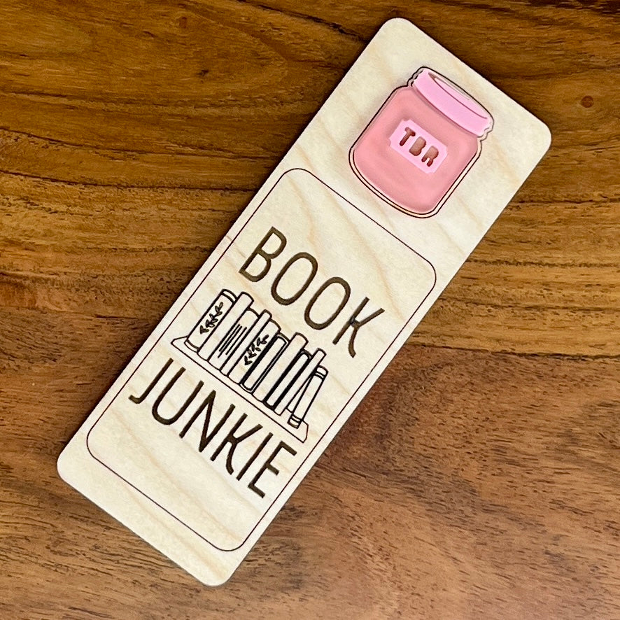 A rectangular wooden book mark with rounded corners, engraved with the words 'BOOK JUNKIE' and a small bookshelf illustration, featuring a pink acrylic TBR jar graphic near the top. The item is photographed on a brown wooden surface.