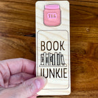 A hand holding a birch wood bookmark with a pink acrylic jar illustration and the laser-engraved words 'BOOK JUNKIE' as well as a engraved bookshelf graphic.