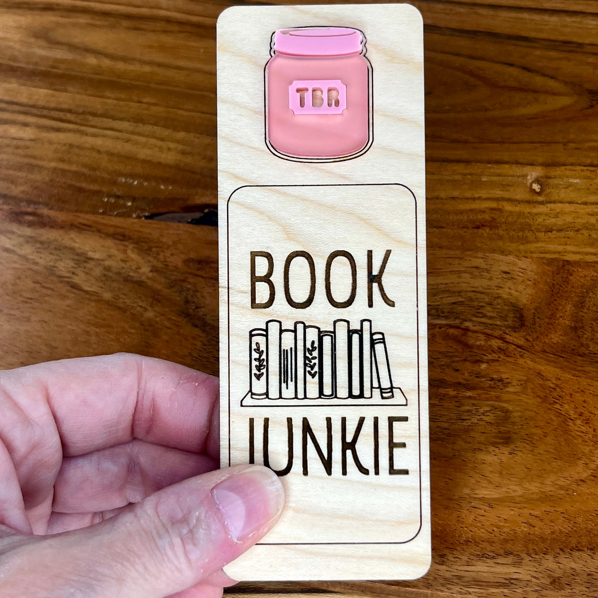 A hand holding a birch wood bookmark with a pink acrylic jar illustration and the laser-engraved words 'BOOK JUNKIE' as well as a engraved bookshelf graphic.