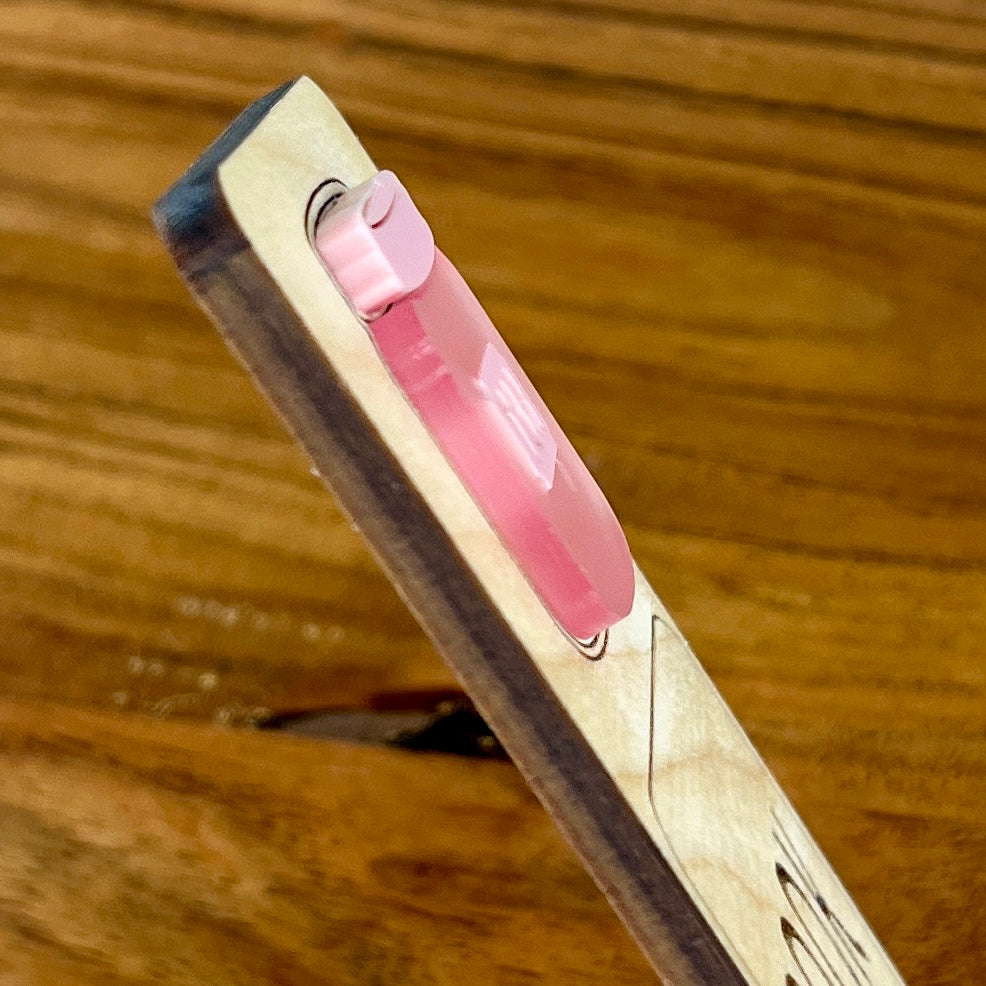 A 3D close-up of a pink translucent  acrylic element layered onto a bookmark, set against a warm wood grain background.