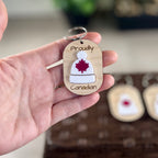 Hand holding Proudly Canadian maple keychain showing white acrylic toque with red maple leaf detail