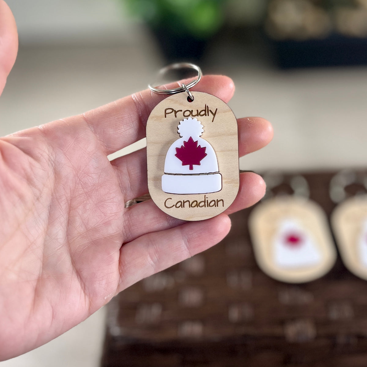 Hand holding Proudly Canadian maple keychain showing white acrylic toque with red maple leaf detail