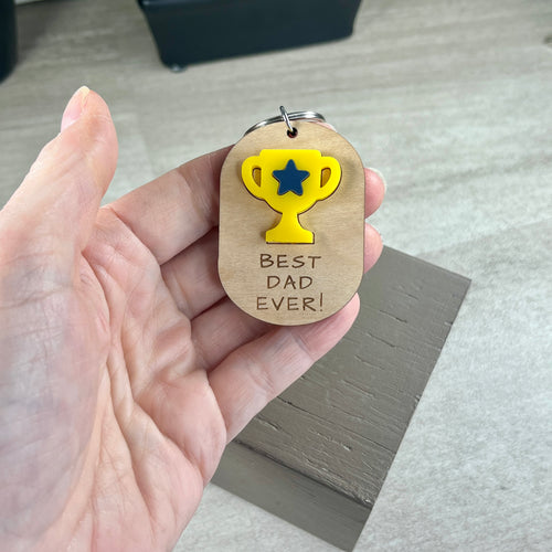 Hand holding Best Dad Ever maple keychain showing yellow acrylic trophy detail