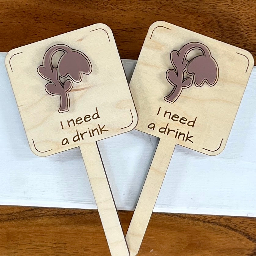 Two I Need a Drink laser-cut maple wood plant stakes with mauve acrylic wilted flower side by side
