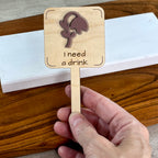 Hand holding an I Need a Drink laser-cut maple wood plant stake with mauve acrylic wilted flower