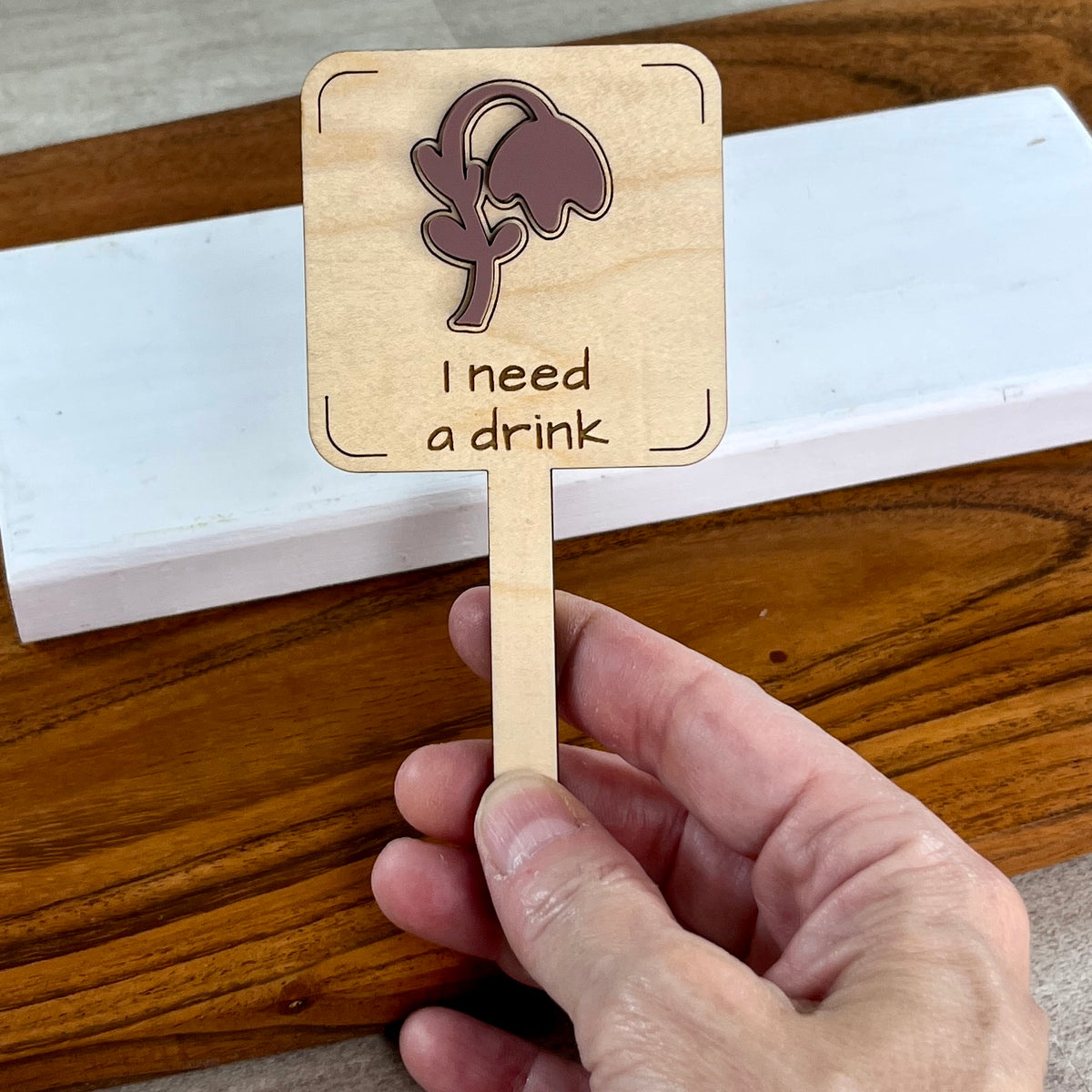 Hand holding an I Need a Drink laser-cut maple wood plant stake with mauve acrylic wilted flower