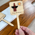 Hand holding a Day Drinker laser-cut maple wood plant stake with wine bottle and sunglasses design