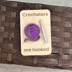 A wooden magnet with the laser-engraved text 'Crocheters are hooked' layered with a purple and lavender acrylic crochet hook and yarn ball, placed on a brown woven basket surface.