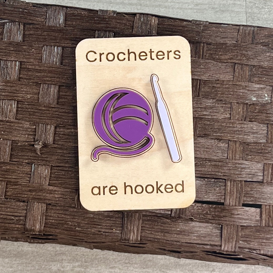 A wooden magnet with the laser-engraved text 'Crocheters are hooked' layered with a purple and lavender acrylic crochet hook and yarn ball, placed on a brown woven basket surface.