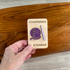 A hand holds a wooden magnet featuring a crocheter-themed acrylic design. The card reads 'Crocheters are hooked' and shows a purple ball of yarn and a lavender crochet hook.