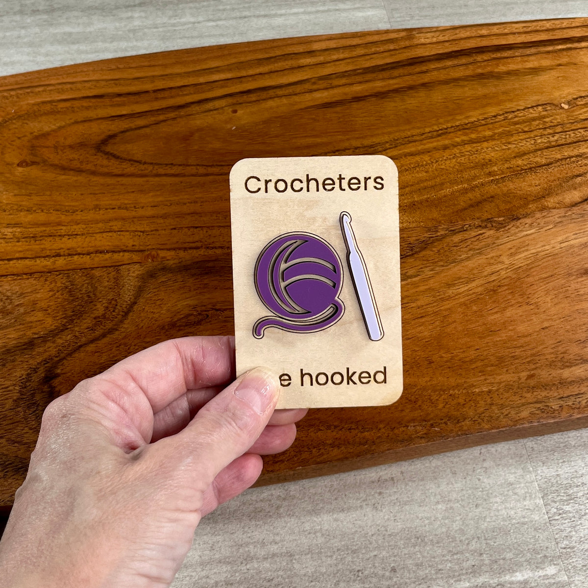 A hand holds a wooden magnet featuring a crocheter-themed acrylic design. The card reads 'Crocheters are hooked' and shows a purple ball of yarn and a lavender crochet hook.