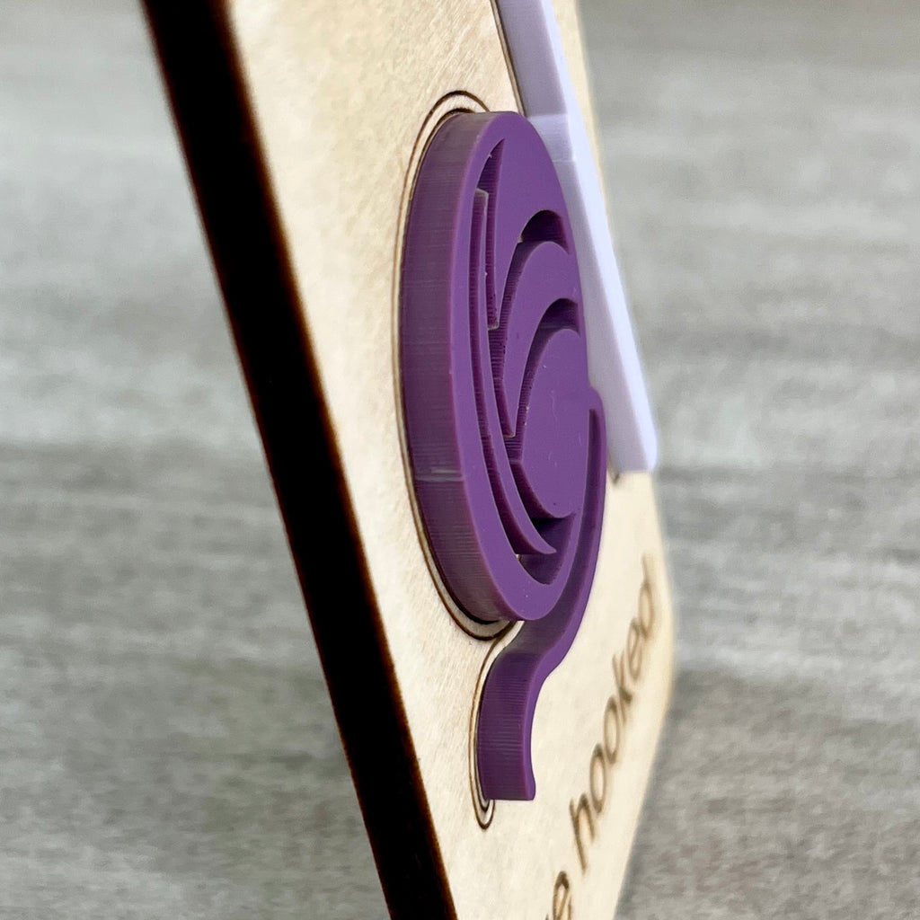 A side view close-up of a wooden magnet with a layered purple acrylic ball of yarn, photographed against a gray textured background highlighting the 3D nature of the product.