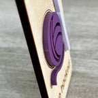 A side view close-up of a wooden magnet with a layered purple acrylic ball of yarn, photographed against a gray textured background highlighting the 3D nature of the product.